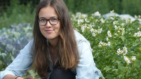 Frau mit langen braunen Haaren, die in einem Garten mit blühenden Pflanzen und weißen Blüten kniet, trägt eine hellblaue Jacke und schwarze Hose.