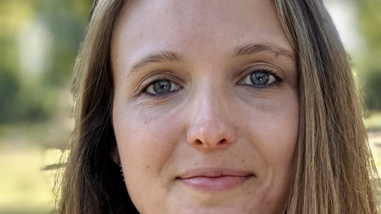 Frau mit langen braunen Haaren, die seitlich über die Schultern fallen, vor einem unscharfen Hintergrund im Freien.