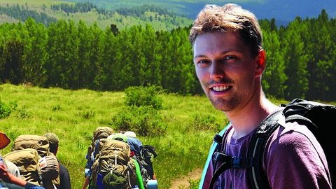Junger Mann mit Rucksack in lila T-Shirt steht auf einer Wiese, im Hintergrund mehrere Wanderer mit Rucksäcken und ein Wald