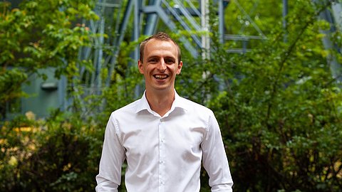 Mann in weißem Hemd steht vor grünen Büschen und einer grauen Stahlstruktur im Hintergrund