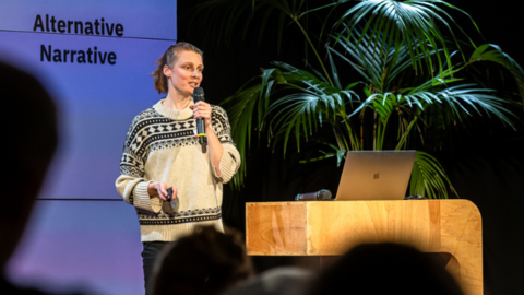 Workshops, Beratung und Formate für strategische Kommunikation von Dr. Linda Walter Frau mit Mikrofon und Präsentationsfernbedienung steht vor Leinwand mit Text 'Alternative Narrative', spricht auf einer Bühne neben Holztisch mit Laptop