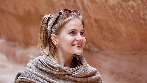 Frau mit hellbraunem Pferdeschwanz, trägt eine graue Strickjacke und eine Sonnenbrille auf dem Kopf, steht vor einer Felswand.