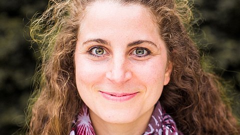 Person mit langen, lockigen braunen Haaren, trägt einen Schal mit lila und weißen Mustern, unscharfer Hintergrund mit grünen Pflanzen