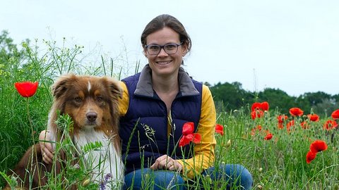 Frau mit dunkelblauer Weste und gelbem Langarmshirt sitzt in einem Mohnblumenfeld und hält eine braune Papiertüte hoch.