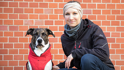 Hund mit braun-weißem Fell trägt rote Weste, sitzt neben Person in schwarzer Jacke und blauer Hose vor roter Backsteinwand