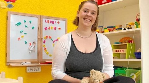 Frau mit weißer Strickjacke und grauem Top sitzt vor gelber Wand mit zwei Magnettafeln, auf denen bunte Buchstaben haften, im Hintergrund Regale mit Spielzeug.