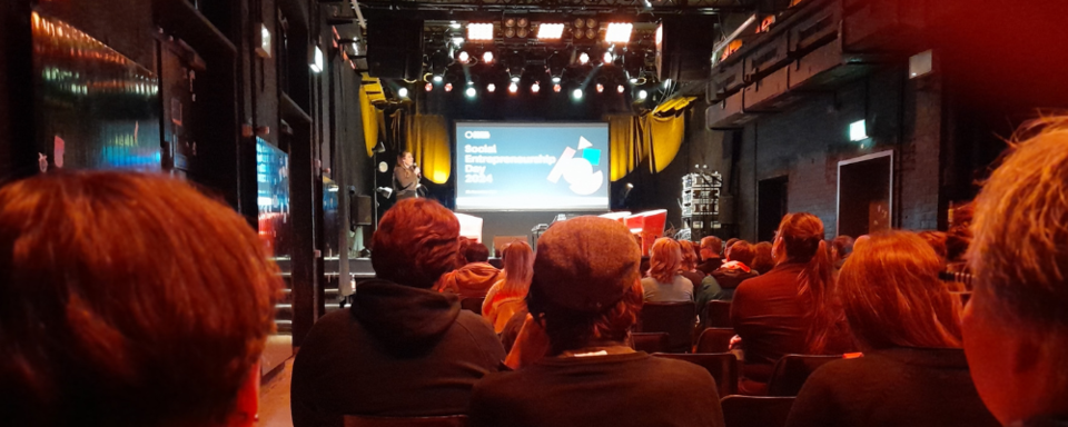 Publikum sitzt in einem Veranstaltungsraum und schaut auf eine Bühne mit einer Präsentation zum Social Entrepreneurship Day 2024