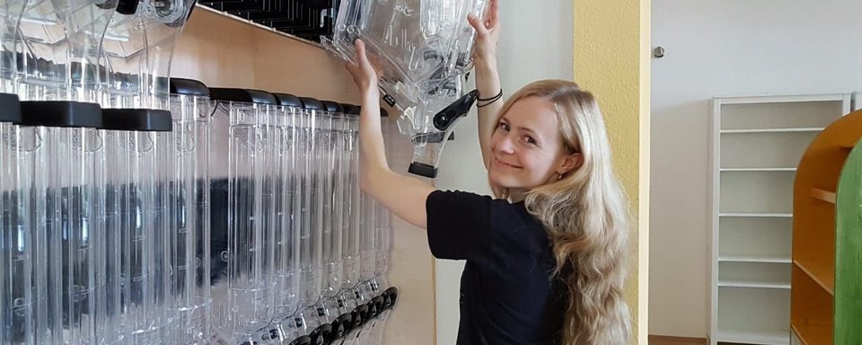 Frau mit langen blonden Haaren, die an einem Regal mit mehreren transparenten Röhren und schwarzen Elementen arbeitet.