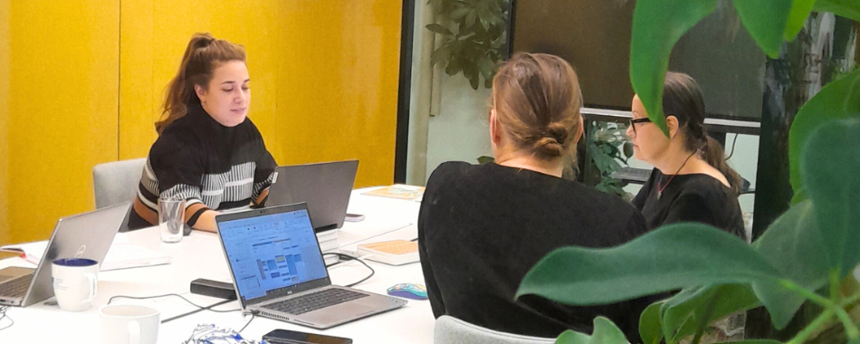 Drei Personen sitzen an einem Tisch in einem Coworking-Space, zwei mit Laptops, umgeben von Pflanzen und gelber Wand