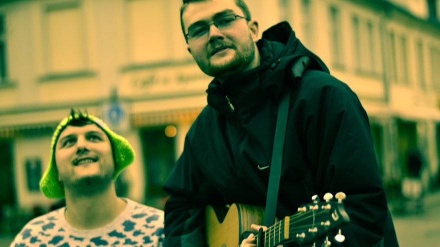 Person in schwarzer Jacke spielt Gitarre auf der Straße, weitere Person mit weißem Shirt mit schwarzem Muster steht daneben