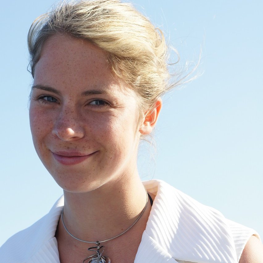 Frau mit hochgestecktem blondem Haar trägt ein weißes, ärmelloses Hemd und eine Kette mit Anhänger vor blauem Himmel