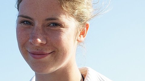 Frau mit hochgestecktem blondem Haar trägt ein weißes, ärmelloses Hemd und eine Kette mit Anhänger vor blauem Himmel