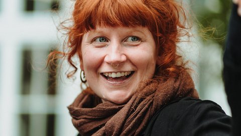 Frau mit roten, lockigen Haaren trägt einen braunen Schal und einen schwarzen Pullover, im Hintergrund unscharfe Fenster