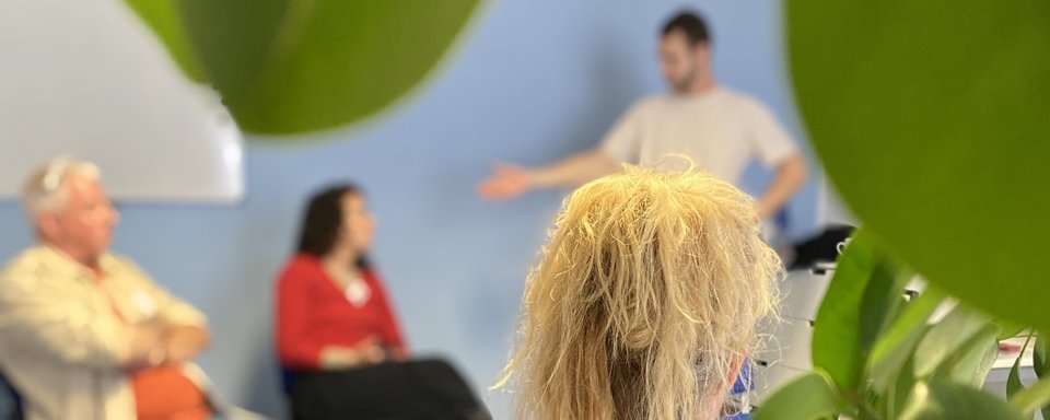 Drei Personen in einem Gespräch, eine steht vor einer blauen Wand und gestikuliert, zwei sitzen und hören zu.