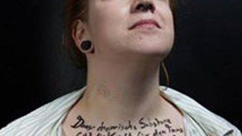 Frau mit roten Haaren, trägt weißes gestreiftes Hemd, sichtbare schwarze Ohrstecker, schwarze Handschrift auf dem Hals