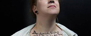 Frau mit roten Haaren, trägt weißes gestreiftes Hemd, sichtbare schwarze Ohrstecker, schwarze Handschrift auf dem Hals