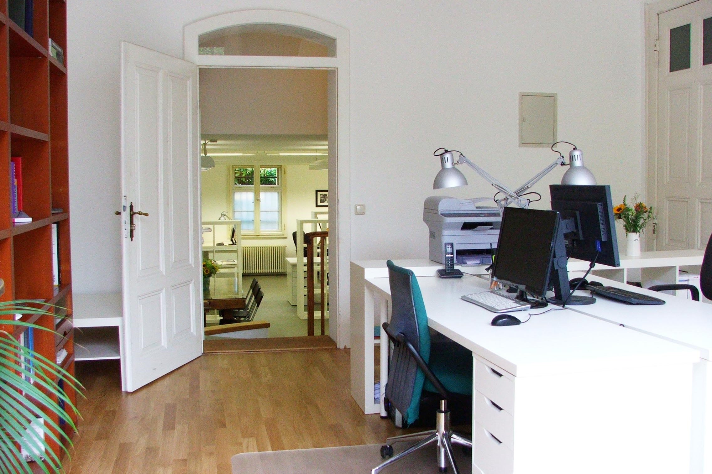 Helles Büro mit weißen Schreibtischen, zwei Computern und Schreibtischlampen, Blick durch offene Tür in weiteren Raum