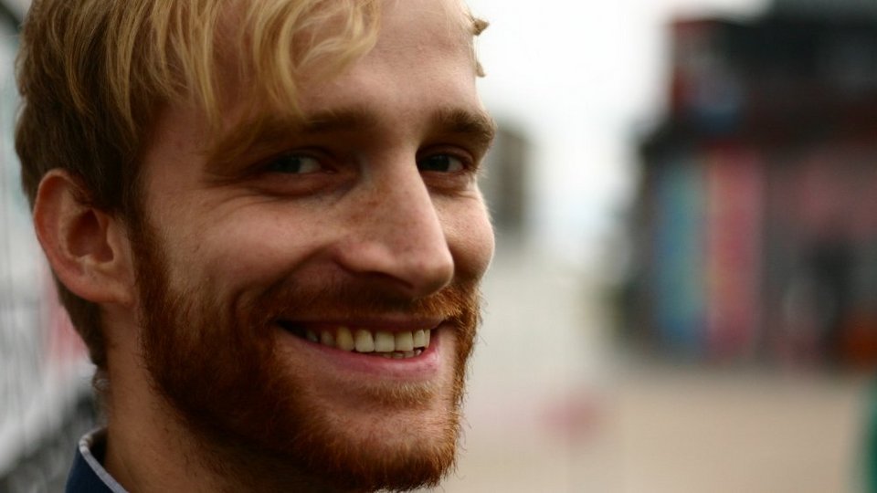 Junger Mann mit kurzen, hellbraunen Haaren, trägt ein dunkelblaues Hemd, im Freien vor unscharfem Hintergrund.