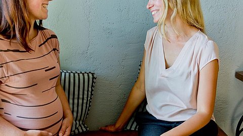 Zwei Frauen sitzen nebeneinander, eine trägt ein gestreiftes, braunes T-Shirt und hält ihren Bauch, die andere trägt ein helles, kurzärmeliges Oberteil.