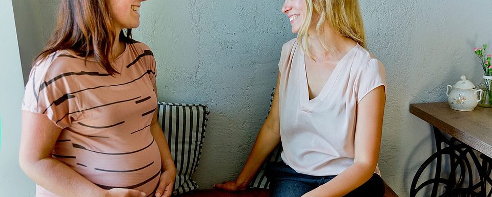 Zwei Frauen sitzen nebeneinander, eine trägt ein gestreiftes, braunes T-Shirt und hält ihren Bauch, die andere trägt ein helles, kurzärmeliges Oberteil.