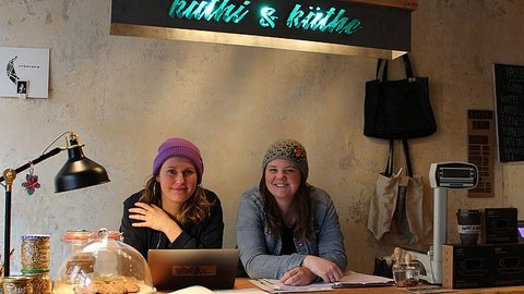 Zwei Frauen sitzen an einem Holztisch mit Laptop, Papieren und einer Lampe in einem gemütlichen Café mit rustikaler Einrichtung.