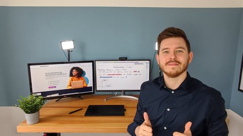 Person in dunklem Hemd sitzt an einem Schreibtisch mit zwei Monitoren, einer zeigt eine Frau, der andere Text, daneben eine grüne Pflanze und eine Wand mit einem Poster