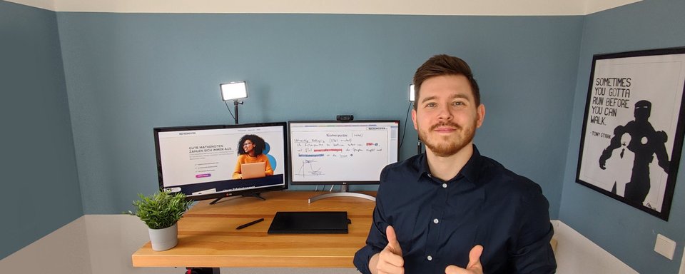 Person in dunklem Hemd sitzt an einem Schreibtisch mit zwei Monitoren, einer zeigt eine Frau, der andere Text, daneben eine grüne Pflanze und eine Wand mit einem Poster