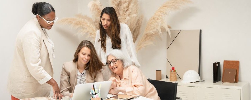 Eine Gruppe von Frauen jedes Alterrs sitzt an einem Tisch und schaut geminsam auf einem Laptop