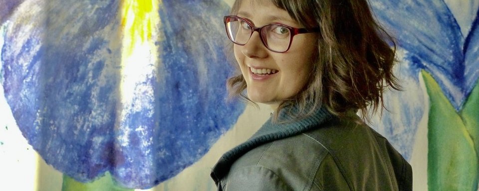 Frau mit braunen, schulterlangen Haaren trägt eine graue Jacke und steht vor einem abstrakten blauen und gelben Gemälde an der Wand