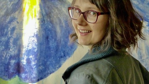 Frau mit braunen, schulterlangen Haaren trägt eine graue Jacke und steht vor einem abstrakten blauen und gelben Gemälde an der Wand