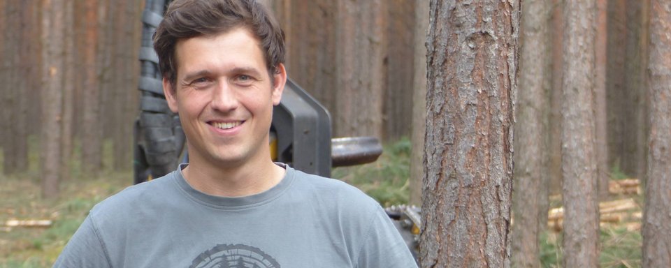 Junger Mann mit kurzem braunem Haar, grauem T-Shirt mit rundem schwarzen Muster, steht vor einem Wald mit vielen hohen Bäumen.