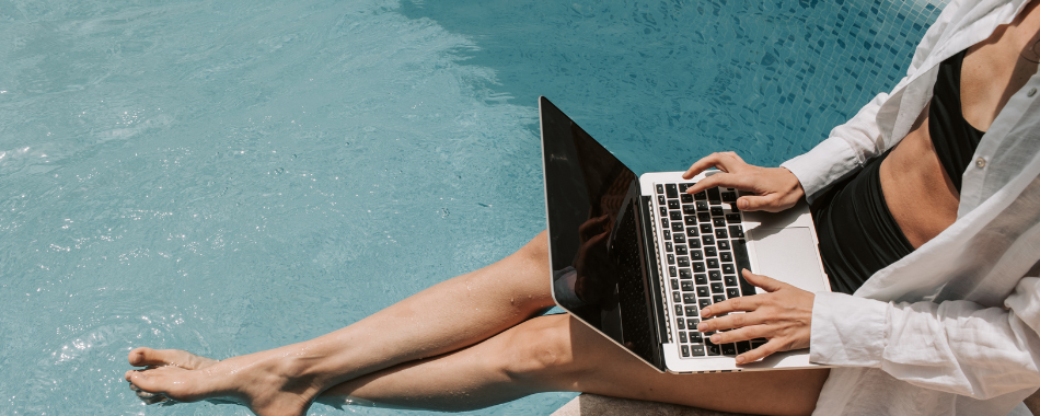 Person sitzt am Poolrand mit Laptop auf dem Schoß, trägt weißen offenen Hemd und schwarzen Badeanzug