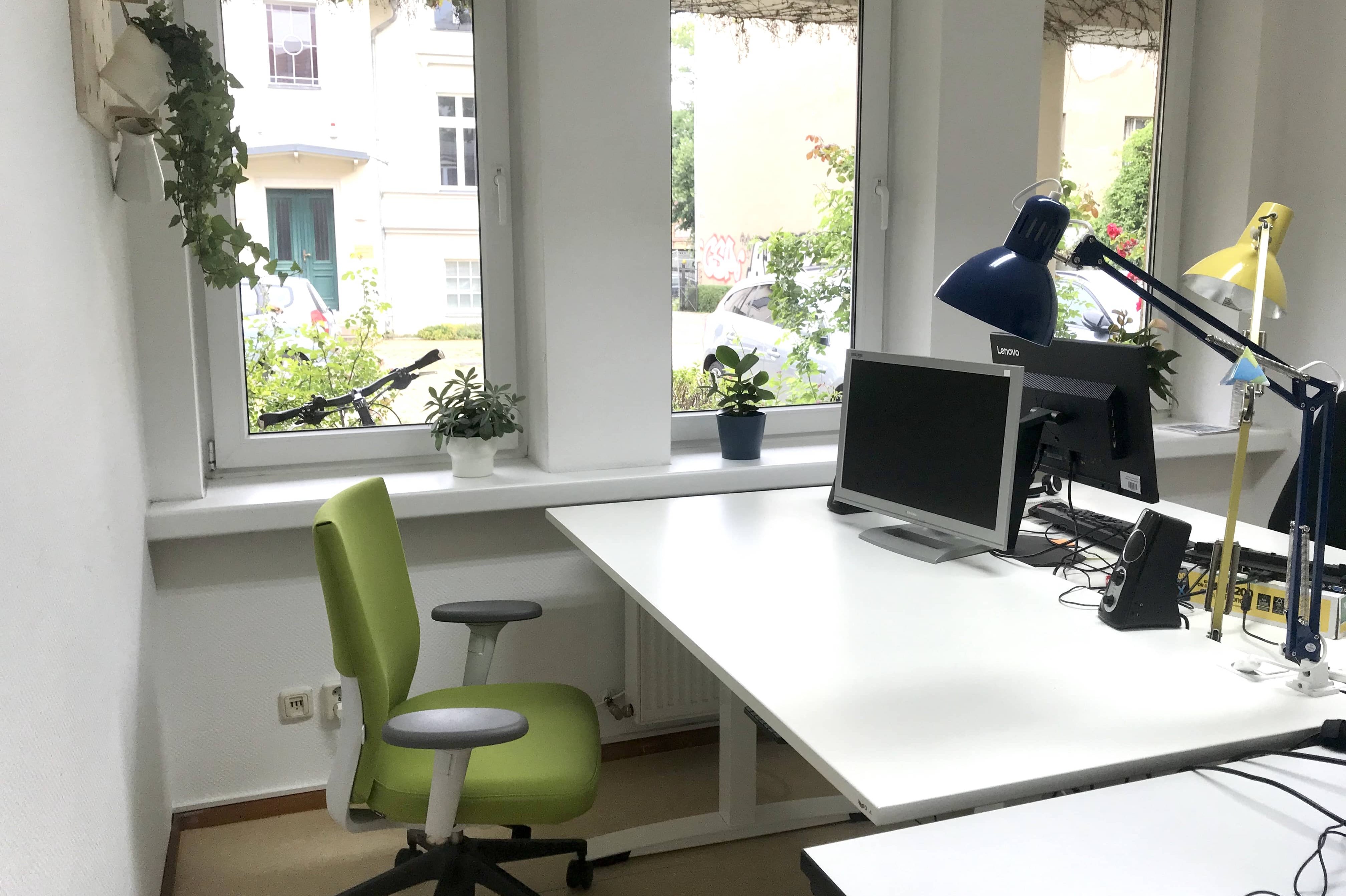 Helles Büro mit drei Fenstern, weißem Eckschreibtisch, grünem Bürostuhl und zwei Schreibtischlampen in Blau und Gelb
