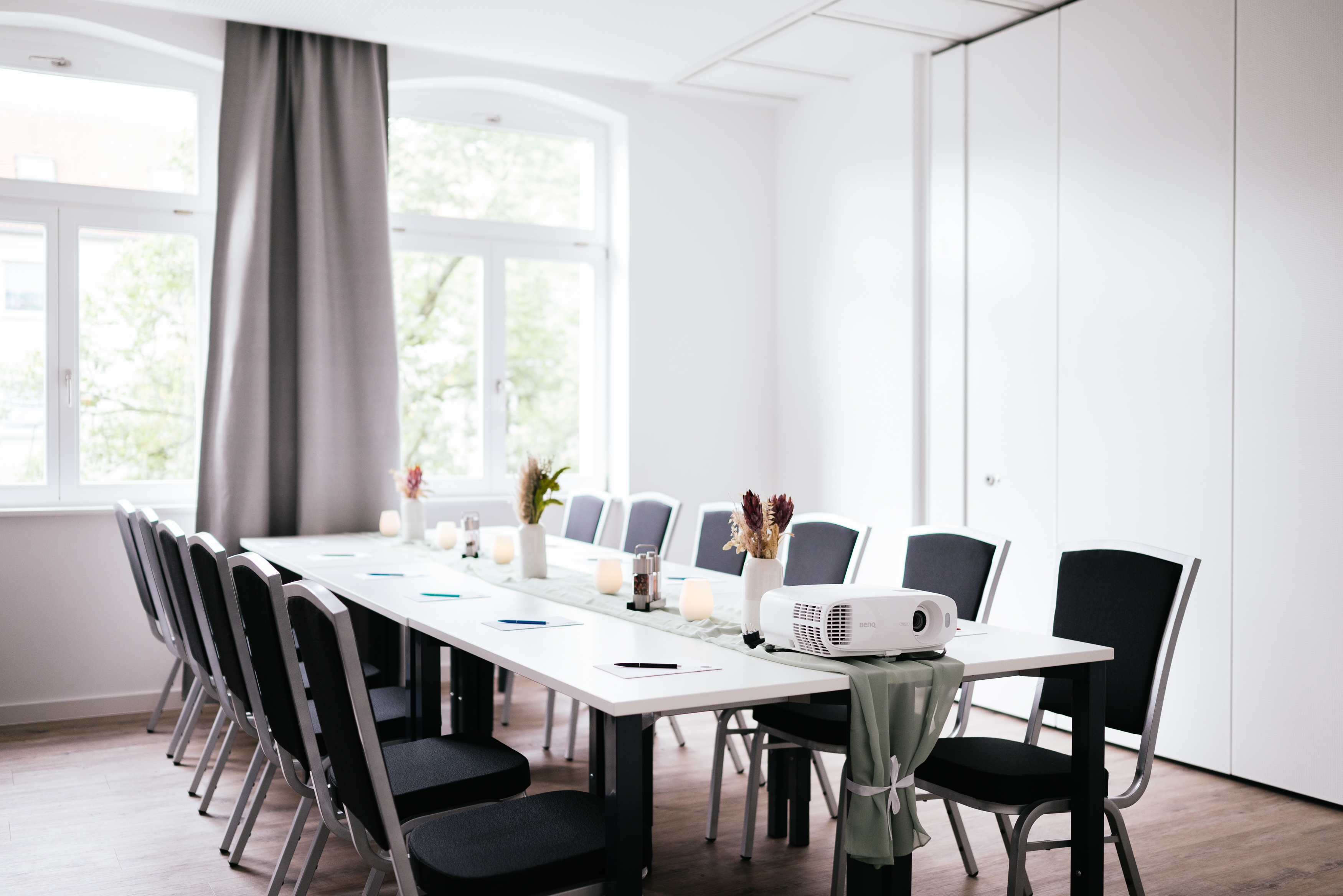 Ein heller Besprechungsraum mit einem langen Tisch, umgeben von schwarzen Stühlen. Auf dem Tisch stehen Kerzen und ein Projektor ist sichtbar