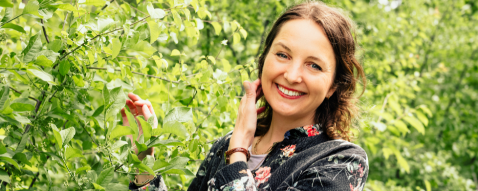 Frau mit lockigem Haar trägt schwarzen Pullover mit Blumenmuster, steht im Grünen und berührt Blätter eines Baums