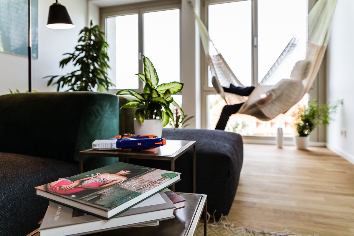 Wohnzimmer mit dunkelgrüner Couch, Pflanzen und Person in Hängematte vor großen Fenstern