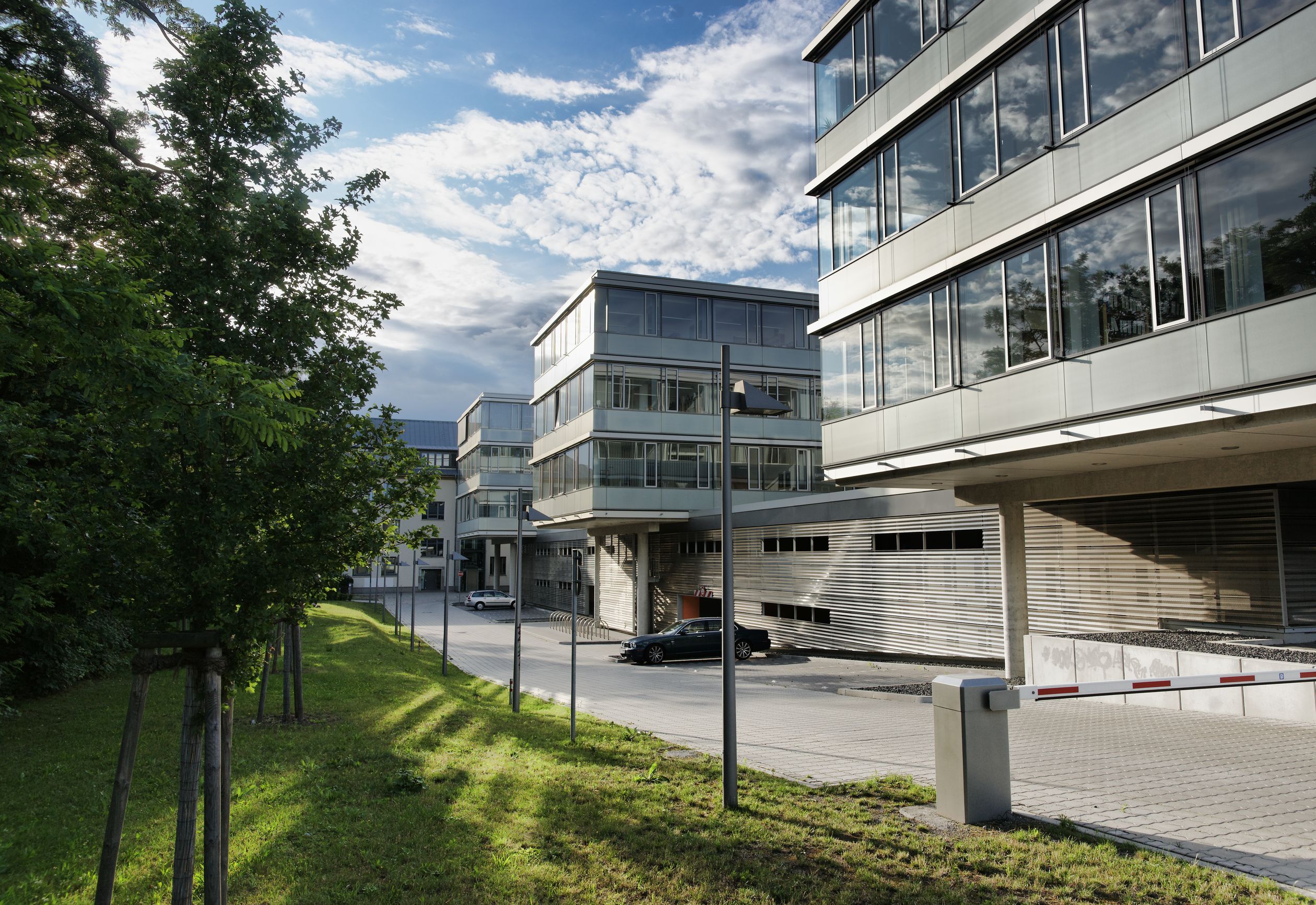 Moderne Bürogebäude mit großen Glasfenstern neben einem grasbewachsenen Weg und Bäumen unter blauem Himmel mit Wolken